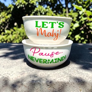 May include: Two white ceramic bowls stacked on top of each other. The top bowl has the words "LET'S Mahj!" in green and orange. The bottom bowl has the words "Pause... NEVERMIND!" in pink and green.
