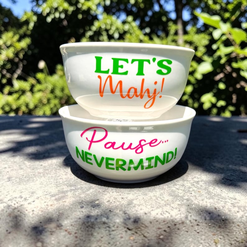 May include: Two white ceramic bowls stacked on top of each other. The top bowl has the words "LET'S Mahj!" in green and orange. The bottom bowl has the words "Pause... NEVERMIND!" in pink and green.