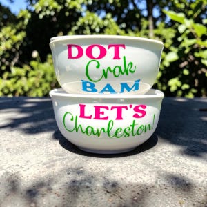 May include: Two white ceramic bowls stacked on top of each other. The top bowl has the words "DOT Crak BAM" in pink, green, and blue. The bottom bowl says "LET'S Charleston!" in pink and green.
