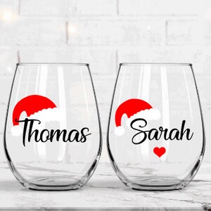 May include: Two clear wine glasses with red and white Santa hats and the names "Thomas" and "Sarah" in black lettering. A red heart is on the glass with "Sarah".