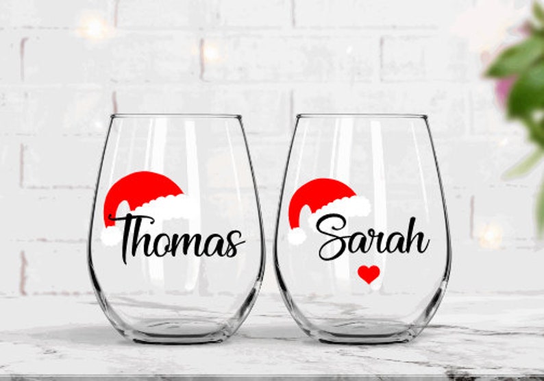 May include: Two clear wine glasses with red and white Santa hats and the names "Thomas" and "Sarah" in black lettering. A red heart is on the glass with "Sarah".