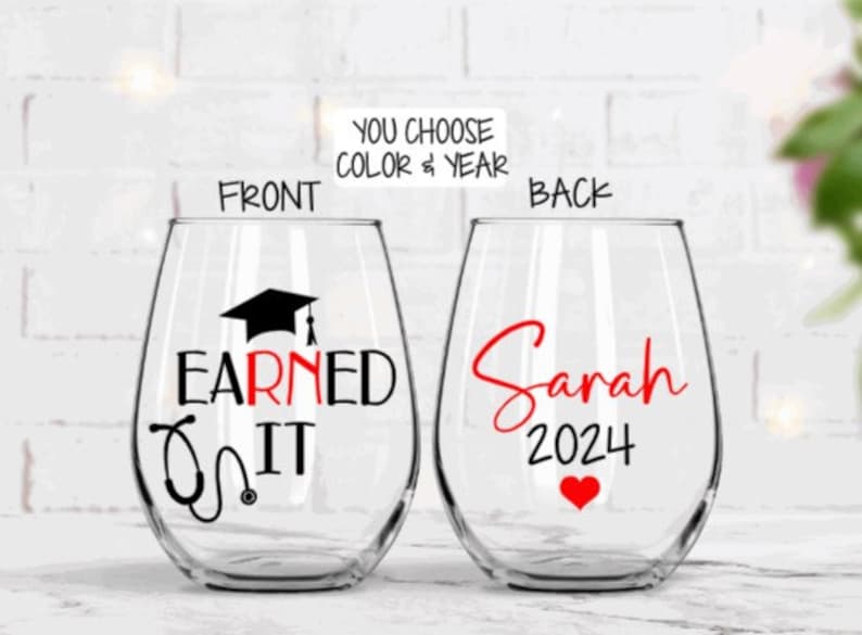 May include: Two stemless wine glasses with a graduation theme. The front glass has a black graduation cap and stethoscope with the text "Earned It". The back glass has the name "Sarah" and the year "2024" in red.