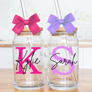 May include: Two glass drinking jars with wooden lids, each featuring a bow on top and a straw. One jar has a pink bow and the name "Kylie" in pink and black. The other jar has a purple bow and the name "Sarah" in purple and black.
