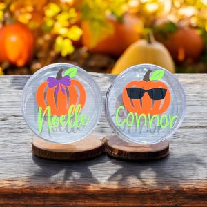 May include: Two clear plastic pumpkin-shaped containers with lids. The container on the left has a purple bow on the pumpkin and the name "Noelle" written in green. The container on the right has sunglasses on the pumpkin and the name "Connor" written in green.