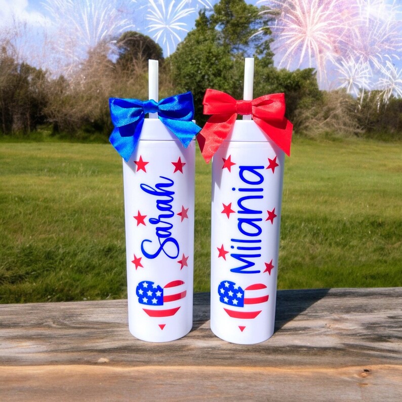 May include: Two white tumblers with red, white, and blue heart designs and stars. The tumblers have the names "Sarah" and "Milania" written on them.