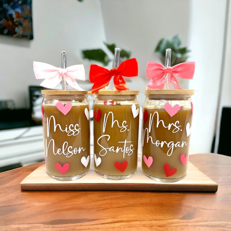 May include: Three glass cups with bamboo lids and straws, decorated with pink and red hearts and personalized with the names "Miss Nelson", "Mrs. Santos", and "Mrs. Morgan".