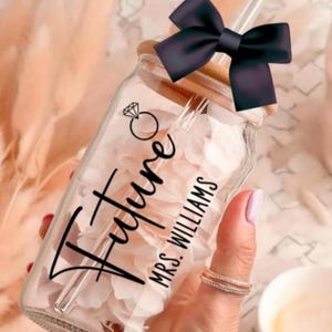 May include: A clear glass jar with a wooden lid and a black bow. The jar has a straw and is decorated with the words "Future MRS. WILLIAMS" and a diamond ring graphic. The jar is filled with pink petals.
