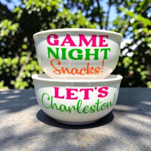 May include: Two white ceramic bowls stacked on top of each other. The top bowl has the words "GAME NIGHT Snacks!" in pink, green, and orange. The bottom bowl says "LET'S Charleston!" in pink and green.
