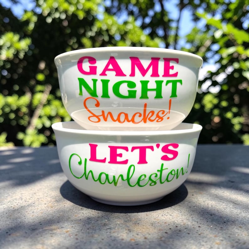 May include: Two white ceramic bowls stacked on top of each other. The top bowl has the words "GAME NIGHT Snacks!" in pink, green, and orange. The bottom bowl says "LET'S Charleston!" in pink and green.