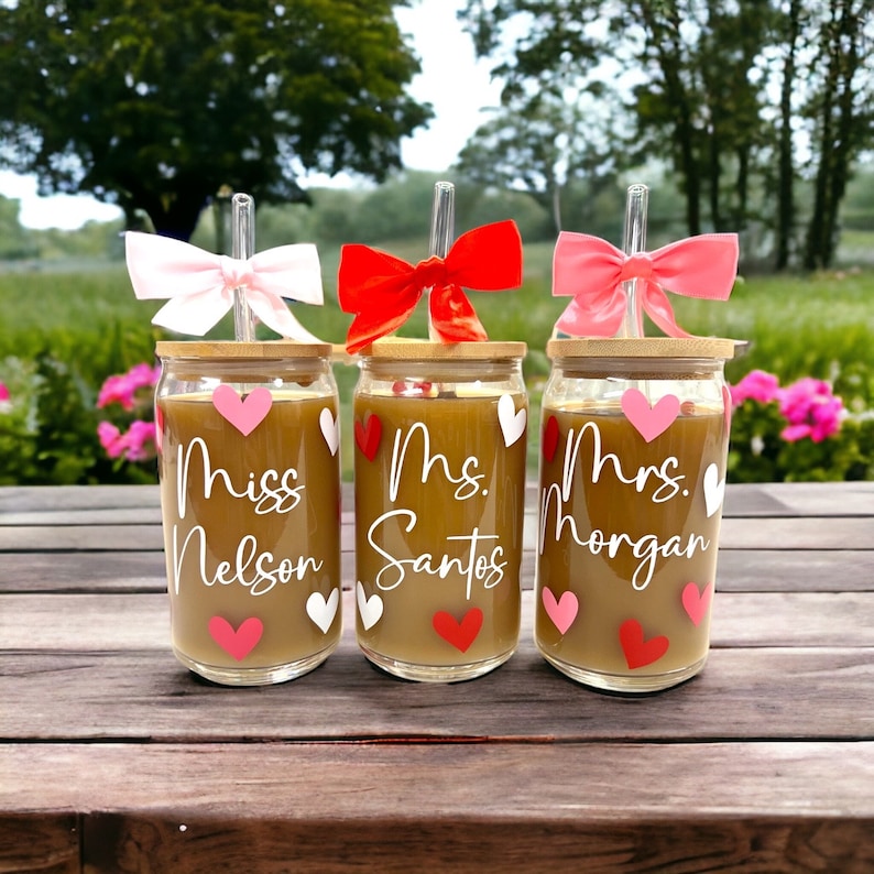 May include: Three glass cups with bamboo lids and straws. Each cup has a different name on it: "Miss Nelson", "Mrs. Santos", and "Mrs. Morgan". The cups are decorated with pink and red hearts.