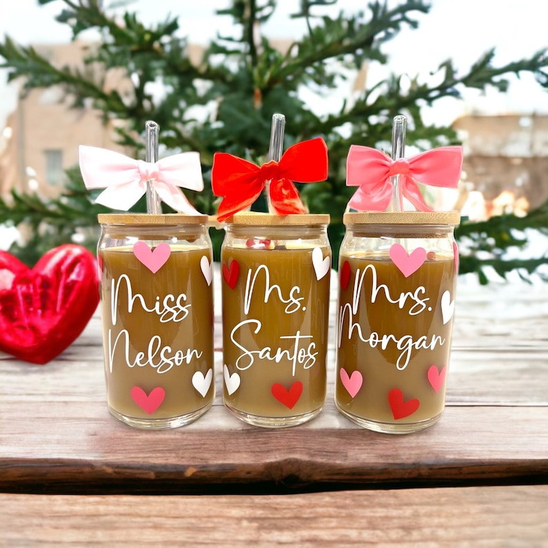 May include: Three glass cups with bamboo lids and straws. Each cup has a different name on it: Miss Nelson, Mrs. Santos, and Mrs. Morgan. The cups are decorated with pink and red hearts.