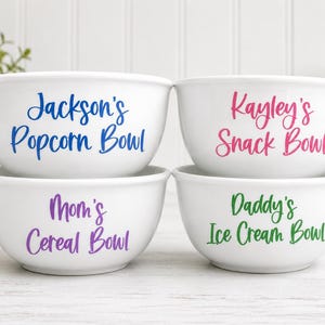 May include: Four white ceramic bowls arranged in a two-by-two grid. The top left bowl says Jackson's Popcorn Bowl in blue. The top right bowl says Kayley's Snack Bowl in pink. The bottom left says Mom's Cereal Bowl in purple. The bottom right says Daddy's Ice Cream Bowl in green.