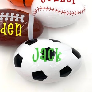 May include: Four plastic Easter eggs decorated with sports themes. The eggs are white with black and white soccer ball design, white with red baseball stitching, brown with white football laces, and orange with black basketball lines. Each egg has a name written on it: Jack, Connor, Den, and an unreadable name.