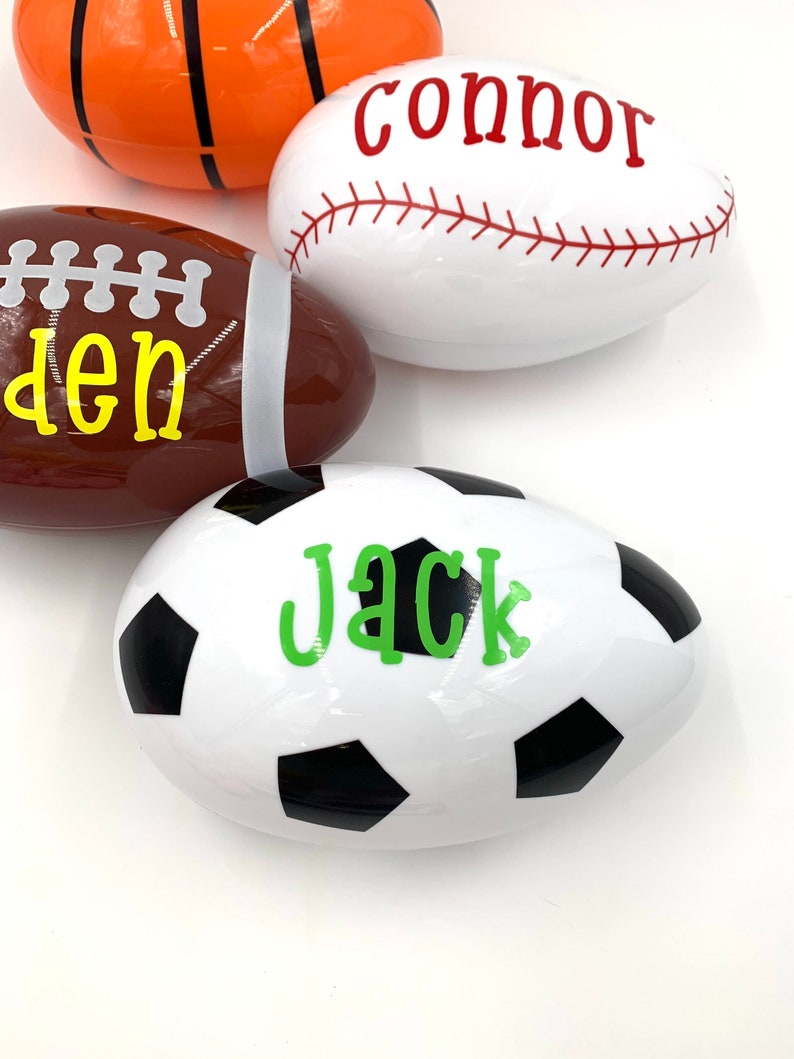 May include: Four plastic Easter eggs decorated with sports themes. The eggs are white with black and white soccer ball design, white with red baseball stitching, brown with white football laces, and orange with black basketball lines. Each egg has a name written on it: Jack, Connor, Den, and an unreadable name.
