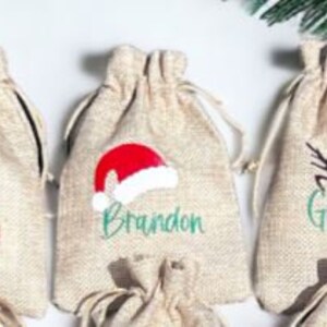 May include: Three small burlap gift bags with personalized Christmas designs. The bags are decorated with a gingerbread man, a Santa hat, and a reindeer. The bags are labeled "Merry", "Brandon", and "Grandma".