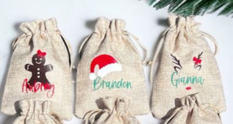 May include: Three small burlap gift bags with personalized Christmas designs. The bags are decorated with a gingerbread man, a Santa hat, and a reindeer. The bags are labeled "Merry", "Brandon", and "Grandma".