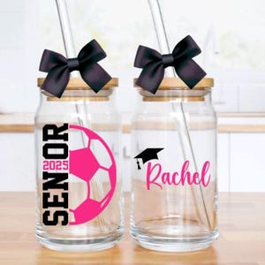 May include: Two glass mason jars with black bows and straws. One jar has a pink soccer ball and the text "Senior 2025". The other jar has a black graduation cap and the name "Rachel".