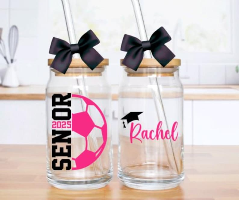 May include: Two glass mason jars with black bows and straws. One jar has a pink soccer ball and the text "Senior 2025". The other jar has a black graduation cap and the name "Rachel".