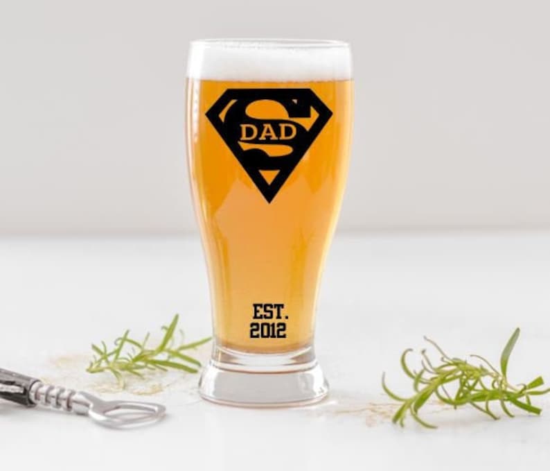 May include: A clear glass beer mug with a Superman logo that says "DAD" and "EST. 2012" printed on the side.