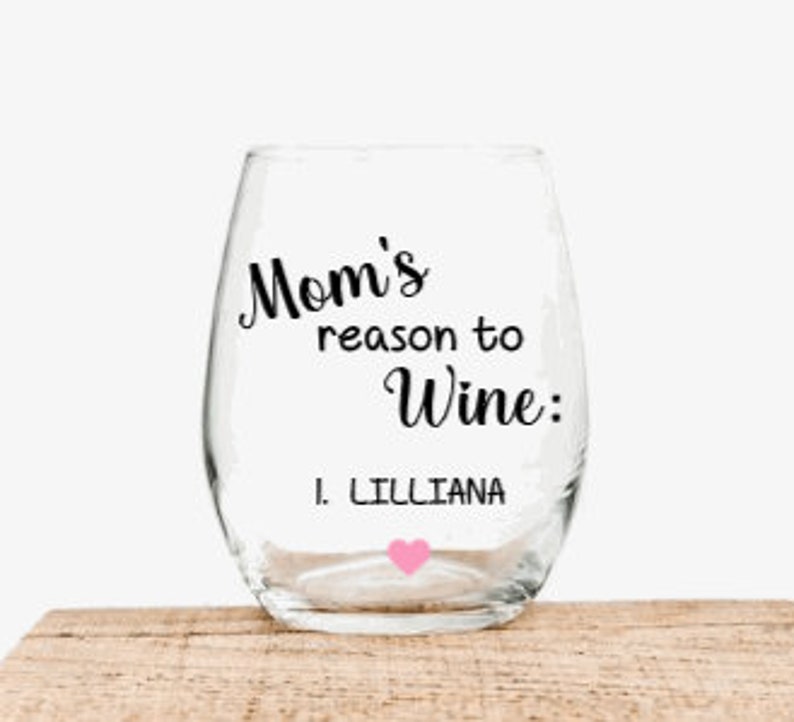 May include: A clear glass wine glass with the text "Mom's reason to wine: 1. Lilliana" in black lettering. A pink heart is below the text.