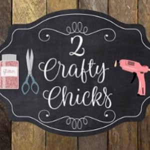 May include: A black chalkboard sign with a white border and the text "2 Crafty Chicks" in white cursive lettering. The sign is decorated with a pair of scissors, a jar of pink glitter, and a pink glue gun.