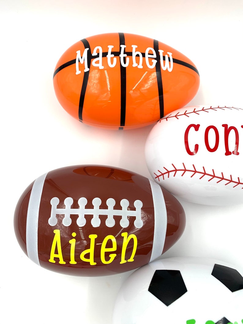 May include: Four plastic Easter eggs decorated with different sports themes. The eggs are white, orange, brown, and white with black accents. The names Matthew, Aiden, and Connor are written on the eggs.