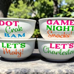 May include: Four white ceramic bowls with colorful text. The top bowl says "DOT Crak BAM" in pink, green, and blue. The next bowl says "GAME NIGHT Snacks!" in pink, green, and orange. The bottom bowls say "LET'S Mahj!" and "LET'S Charleston!"