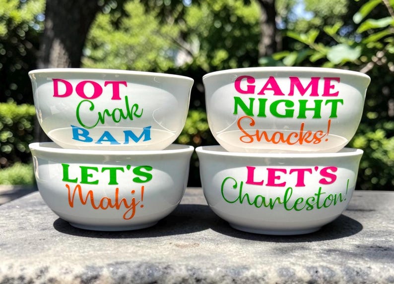 May include: Four white ceramic bowls with colorful text. The top bowl says "DOT Crak BAM" in pink, green, and blue. The next bowl says "GAME NIGHT Snacks!" in pink, green, and orange. The bottom bowls say "LET'S Mahj!" and "LET'S Charleston!"
