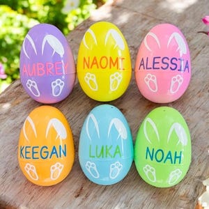 May include: Six plastic Easter eggs decorated with bunny ears and footprints. Each egg is a different color and has a name written on it: Aubrey, Naomi, Alessia, Keegan, Luka, and Noah.