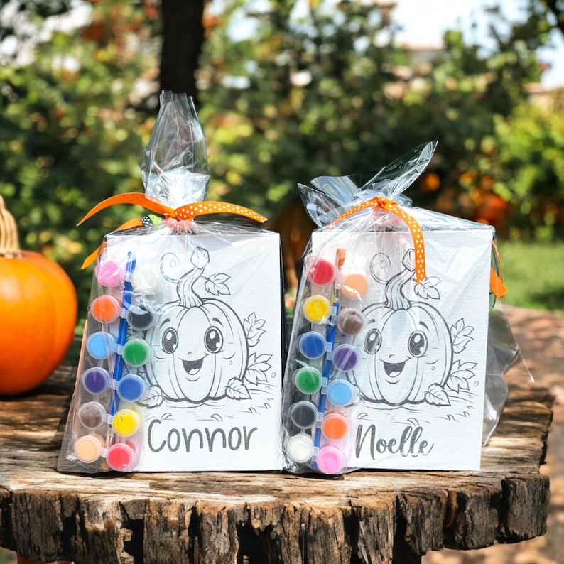 May include: Two pumpkin-themed paint-your-own kits, each with a small canvas, paintbrushes, and a set of paints. The canvases feature a black and white outline of a smiling pumpkin with the names "Connor" and "Noelle" written below.