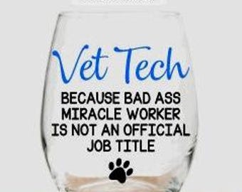 May include: A clear stemless wine glass featuring blue text that reads Vet Tech and black text that reads BECAUSE BAD ASS MIRACLE WORKER IS NOT AN OFFICIAL JOB TITLE with a black paw print icon. Above the glass is text CHOOSE COLOR OPTION TO ADD NAME TO BACK.
