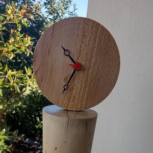 May include: A handcrafted wooden clock with a round face and black hands, featuring a red second hand. The clock is mounted on a cylindrical wooden base with natural wood grain and imperfections. The clock's diameter is approximately 4 inches.