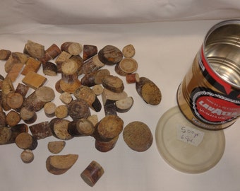 May include: A collection of small, brown wooden pieces, some round and some rectangular, scattered on a white surface. A silver can with a red and gold label that reads "Lavazza Qualita Oro" is next to the wooden pieces. The can is open and empty. The lid of the can is white with the text "500gr 694" written on it.
