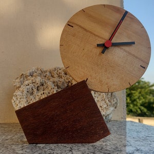 Pode incluir: Um relógio de madeira com um ponteiro dos segundos vermelho e ponteiros das horas e dos minutos pretos. O relógio está montado em uma base de madeira com uma base de pedra áspera de cor marrom claro.