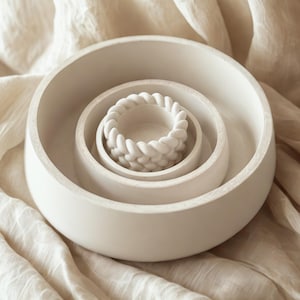 Puede incluir: Un conjunto de tres cuencos de cerámica blancos, circulares y anidados. El cuenco más pequeño tiene un diseño trenzado decorativo en el borde. Los cuencos están colocados sobre una tela beige texturizada.