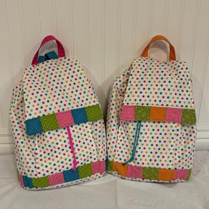 Può includere: Due zaini in tessuto a pois colorati su sfondo bianco. Uno ha finiture rosa e blu, l'altro finiture arancioni e verdi. Ogni zaino ha un manico di colore diverso. Gli zaini sono in tessuto.