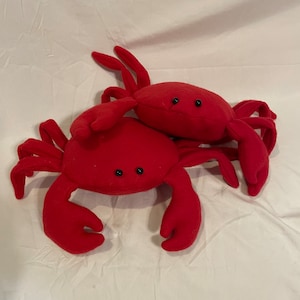 May include: Two plush red crab toys with black button eyes and multiple legs. The crabs are made of a soft material and are resting on a white surface. The crabs are a fun, whimsical design.