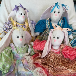May include: Four handmade bunny dolls with white faces, pink noses, and long ears. Each doll wears a colorful dress with lace trim and a matching bow. The dresses feature floral and patterned designs in shades of pink, yellow, blue, and green.