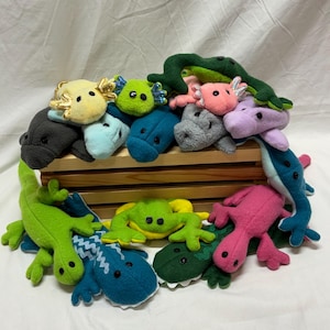 May include: A collection of colorful plush toy amphibians and reptiles, including frogs, axolotls, and lizards. The toys are in various shades of green, blue, pink, yellow, and gray. They are arranged on a wooden crate.