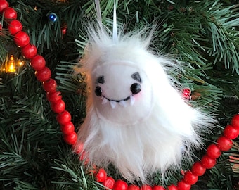 Puede incluir: Adorno navideño de yeti blanco y esponjoso con una cara sonriente, ojos negros y pequeños colmillos. El adorno cuelga de una cinta blanca sobre un fondo de un árbol de Navidad decorado y una guirnalda de cuentas rojas.