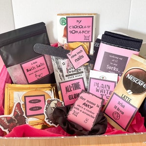 May include: A pink gift box filled with various items, including bath salts, a face mask, a chocolate bar, socks, and a coffee packet. The box also includes several pink and black stickers with text such as "My Chocolate Romance", "Nine Inch Nails", "All Time Glow", "Plain White T's", and "Her name is Amy, she's a rocker with a scrunchie".