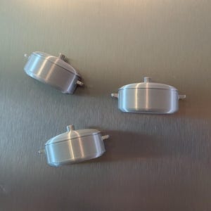 May include: Three small, silver-colored pot-shaped magnets are attached to a stainless steel refrigerator door. Each magnet has a lid and two small handles. The magnets are arranged in a triangular pattern.