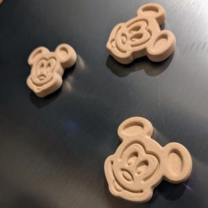 May include: Three wooden Mickey Mouse-shaped cutouts. The cutouts are light brown and have a three-dimensional design. The Mickey Mouse cutouts are placed on a metallic surface. The cutouts are likely for crafting or decoration.