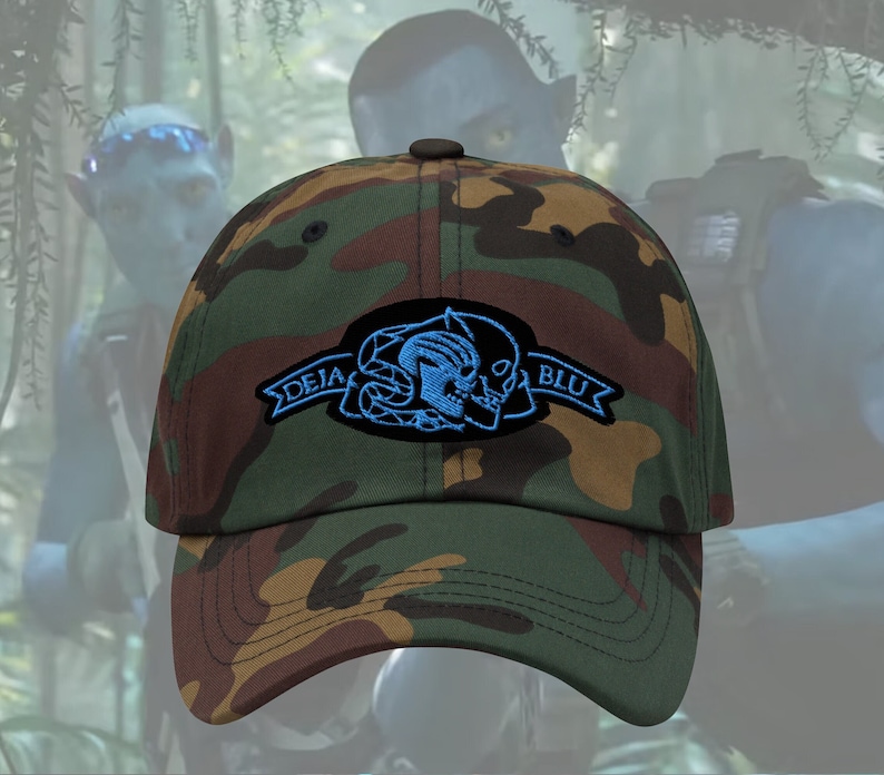 Puede incluir: Gorra de b&eacute;isbol de camuflaje con un parche negro de calavera y huesos cruzados con detalles azules y el texto "DEJA BLU" en una pancarta.