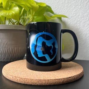 May include: Black ceramic mug featuring a blue and white graphic of two faces in profile, facing each other. The faces are silhouetted against a blue circle with a white outline.