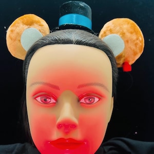 May include: A brown and gray headband with bear ears and a blue and black top hat. The headband is worn on a mannequin with red-toned lighting.