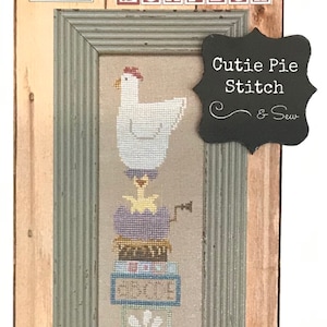 May include: A framed cross-stitch design titled "Cutie Pie Stitch & Sew" featuring a white chicken, a stack of colorful blocks with words like "Spring" and "For You", and a bee. The frame is a light green wood with a natural wood border.