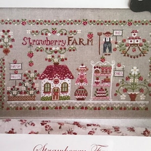 May include: A cross-stitch pattern featuring a charming strawberry farm scene. The pattern includes a small house, a strawberry stand, and a farmer. The text "Strawberry FARM" is stitched in red and white. The stitch count is 250 x 150.
