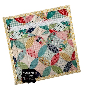 May include: A quilted fabric pouch with a zipper closure and a white lace trim. The pouch is decorated with a patchwork design of colorful circles and has a black label that says "Cutie Pie Stitch & Sew".