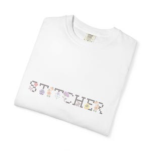May include: White t-shirt with the word "STITCHER" in a cross-stitch style, featuring floral and heart accents. The design incorporates pastel colors and black stitching, creating a handcrafted aesthetic. The shirt is folded.